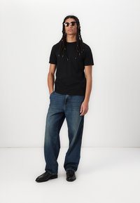 Svart bomull t-skjorte, løse blå denimjeans og svarte sko. Modellen har på seg solbriller; håret er stylet i lange fletter.