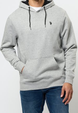 Man draagt een lichtgrijze hoodie met zwarte trekkoorden en een klein donker logo op de borst, gecombineerd met een blauwe spijkerbroek, één hand in de zak.