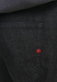 Zbliżenie czarnej tkaniny dżinsowej z szczegółowym przeszyciem oraz małym czerwonym, okrągłym haftem w pobliżu krawędzi kieszeni.