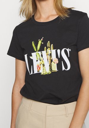 Frau trägt ein schwarzes Levi's T-Shirt mit weißem Logo, teilweise ausgefüllt mit grünen und pinkfarben blühenden Kakteen, kombiniert mit beigen Hosen.