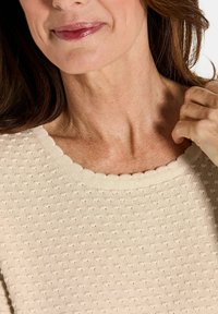 Nahaufnahme des unteren Gesichts einer Frau, ihres Halses und ihrer Hand, die den gewellten Ausschnitt ihres beigefarbenen strukturierten Pullovers anpasst.