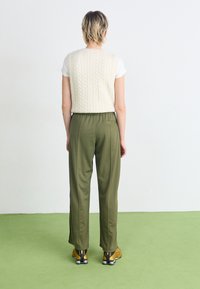 Ljust beige kabelstickad väst över en vit t-shirt, kombinerat med olivgröna byxor. Outfiten har sidofickor och svarta och guldskor.