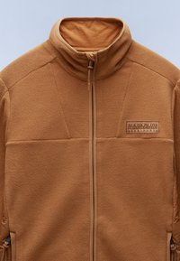 Braune Fleecejacke mit hohem Kragen, Frontreißverschluss und einem Logo-Patch auf der Brust. Strukturierter Stoff mit kontrastierenden beigen Akzenten.