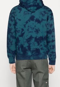 Sweat à capuche en motif tie-dye bleu sarcelle foncé et marine. Il présente une poche kangourou, des poignets et un ourlet côtelés, fabriqué en coton doux.