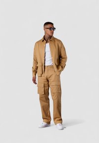 Tan cotton blend jacket with snap buttons and matching cargo pants featuring multiple pockets, paired with a white tank top and sneakers.