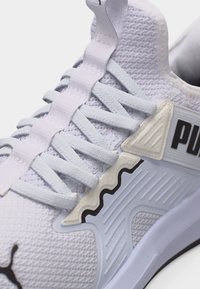 Κοντινό πλάνο λευκού αθλητικού παπουτσιού Puma που δείχνει υφασμάτινη υφή, κορδόνια, πλαστικό στήριγμα κορδονιών και μαύρο λογότυπο Puma στο πλάι.