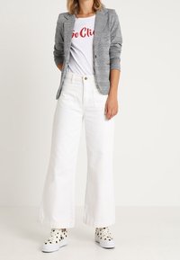 Šedé kostkované sako přes bílé tričko s červeným nápisem, doplněné širokými bílými kalhotami a černými teniskami s puntíky.