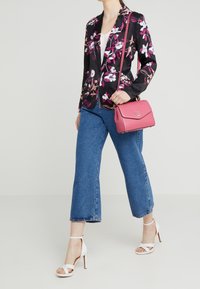Nainen, joka wear kukkakuvioista mustaa bleiseriä, sinisiä cropped-farkkuja, valkoisia korkokenkiä ja kantaa vaaleanpunaista olkalaukkua.
