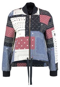 Veste bomber matelassée présentant un mélange de motifs géométriques en bordeaux, marine, blanc et bleu. Comprend une fermeture éclair à l'avant et des poignets côtelés.