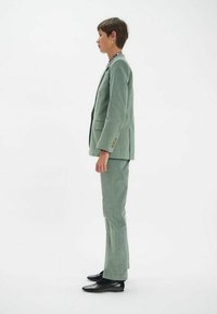 Un costume vert clair en tissu texturé, comprenant un blazer ajusté avec deux poches et un pantalon assorti, associé à des chaussures noires.