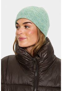 Žena v světlém zeleném pleteném kulichu a tmavě hnědé péřové bundě s vysokým límcem, hledící vlevo a mírně se usmívající.