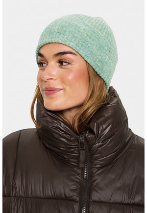 Ženska nosi svetlo zeleno pleteno kapo in temno rjavo puhovko z visokim ovratnikom, gleda levo in se rahlo smehlja.