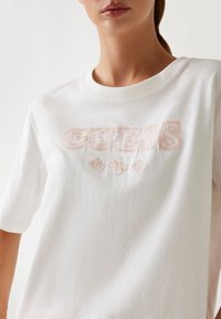 T-shirt in cotone bianco con la parola "GUESS" ricamata in rosa chiaro. Maniche corte e scollatura tonda con una texture liscia.