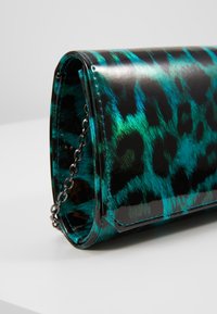 Clutch leopard print lucido turchese e nero con tracolla in argento, appoggiato su una superficie bianca contro uno sfondo grigio chiaro.