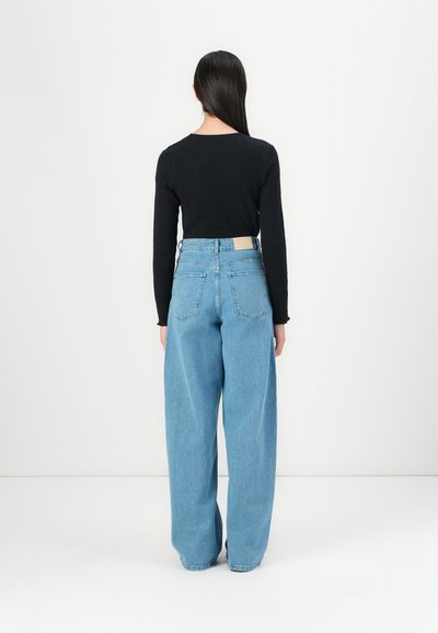 Zwarte geribbelde blouse met lange mouwen, gecombineerd met lichtblauwe wijduitlopende jeans met een beige patch op de tailleband en twee achterzakken.