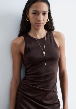 Vrouw met lang donker haar draagt een mouwloze bruine jurk, gouden oorbellen in de vorm van ringen en een gouden hangersketting tegen een witte achtergrond.