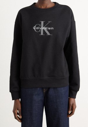 Personne portant un sweat-shirt noir Calvin Klein avec un grand logo "cK" et le texte blanc "Calvin Klein", associé à un jean bleu foncé.