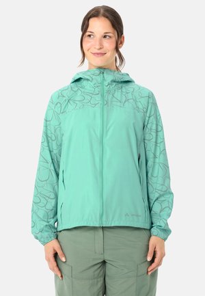 Vrouw draagt een teal kleurige jas met rits en bloemenpatronen op de mouwen en kap, gecombineerd met groene cargobroek, kijkt lichtjes opzij.