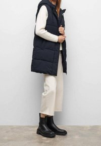 Černá zateplená vesta s kapucí, zapínáním na zip vpředu a bočními kapsami, nošená přes bílý svetr a krémové kalhoty. Černé kotníkové boty.