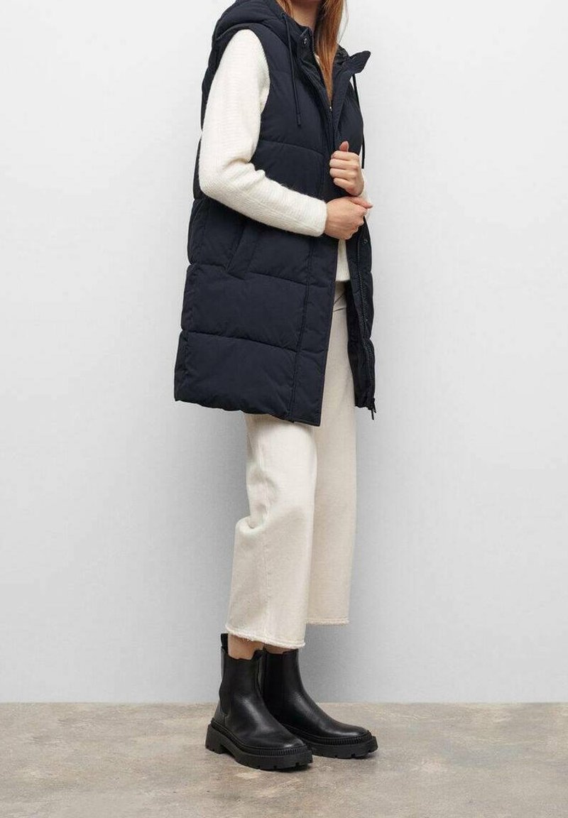 Černá zateplená vesta s kapucí, zapínáním na zip vpředu a bočními kapsami, nošená přes bílý svetr a krémové kalhoty. Černé kotníkové boty.