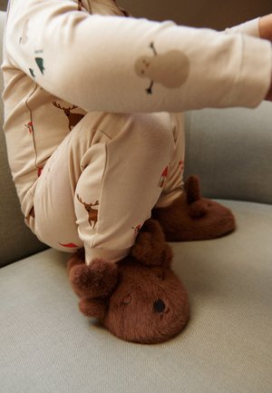 Enfant portant un pyjama beige avec des imprimés d’animaux et des chaussons ours marron duveteux assis sur un canapé gris clair.