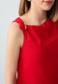 Top rosso senza maniche realizzato in tessuto liscio, con dettaglio spalla attorcigliato e un accento metallico. Design semplice ed elegante.