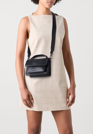 Bolso de cuero negro con estampado de cocodrilo, asa superior y correa de hombro ajustable, de forma rectangular, presentado sobre un vestido de cuadros claros.