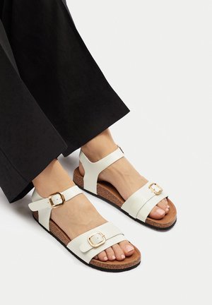 Białe sandały ze skóry z dwoma paskami, złotymi klamrami i korkową wkładką. Podeszwa jest czarna, wykończona teksturą.