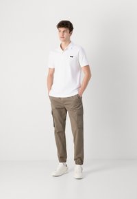 Biała koszulka polo z małym czarnym logo, zestawiona z jasnobrązowymi spodniami cargo z kieszeniami, elastycznymi mankietami i białymi sneakersami.