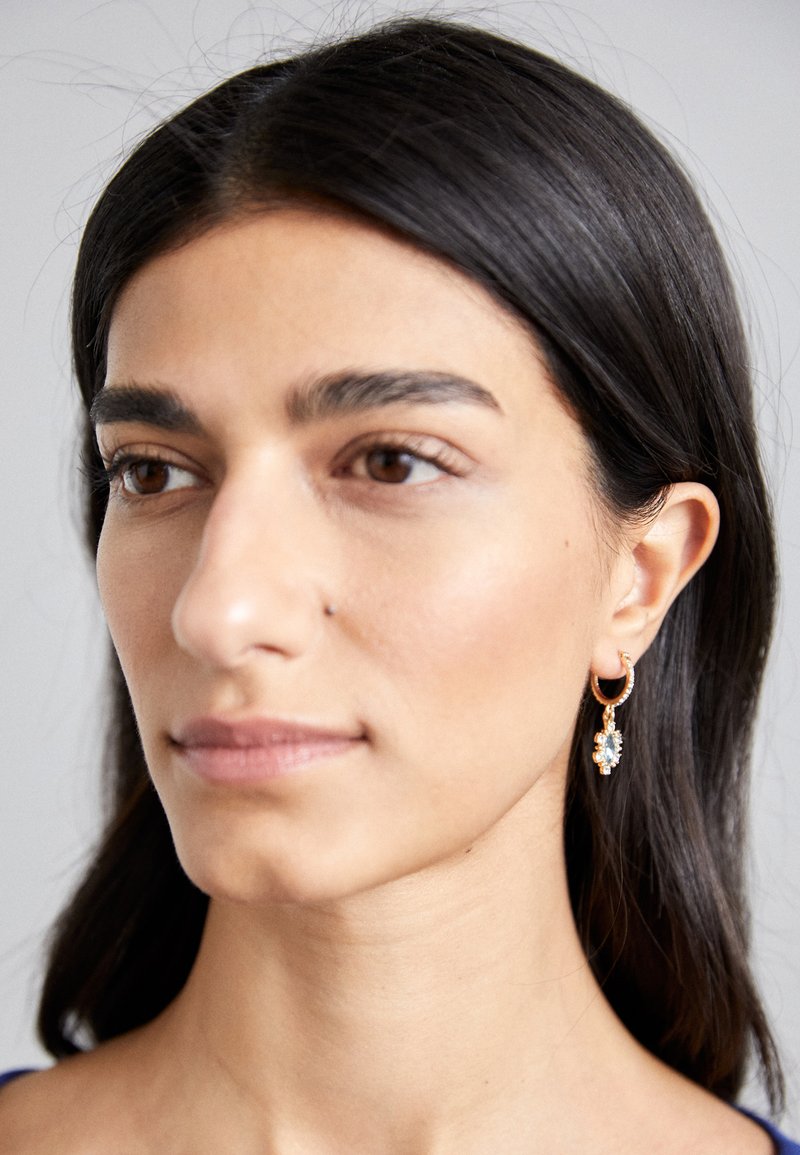 Orecchini a cerchio in oro con design floreale, dotati di accenti in pietra preziosa trasparente. Texture liscia e forma delicata.