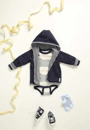 Marineblå spædbarnshættetrøje lagdelt over en stribet body, parret med blå sokker og sorte sneakers. Har blødt stof og trykknapper.