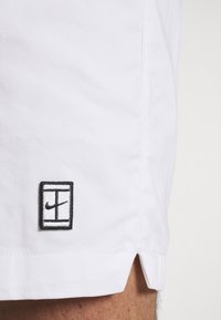Tissu blanc avec une petite étiquette rectangulaire noire et blanche près de l'ourlet, montrant une fente fendue et une partie de la jambe d'une personne.