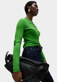 Maglione polo verde a coste con bottoni, abbinato a jeans scuri. Il modello tiene una grande borsa nera intrecciata con una tracolla in pelle.
