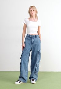 Jeans cargo évasés en denim bleu clair, dotés de plusieurs poches latérales. Associés avec un crop top blanc ajusté et des baskets basses.