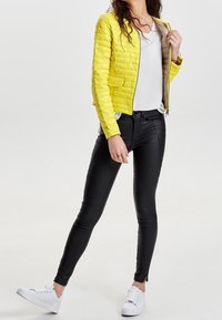 Yellow quilted jacket with a slim fit, two front pockets, paired with black leather-like pants and white sneakers.