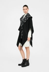 Poncho nero con interno grigio a motivo, dotato di un colletto oversize e chiusura con cravatta. Il modello indossa un vestito crema e stivaletti neri.