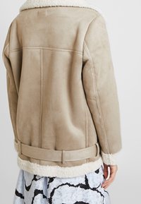 Beige suede jacket with a soft, white shearling collar and cuffs. The design features a belted waist and visible stitching details.