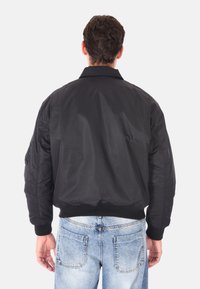 Giubbotto bomber nero con colletto e polsini a costine, materiale liscio e vestibilità rilassata, visto da dietro, abbinato a jeans in denim chiaro.