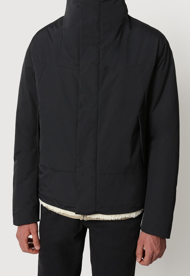 Homme portant une veste noire zippée avec un col montant et une doublure en polaire blanche visible à l'ourlet, associée à un pantalon noir.