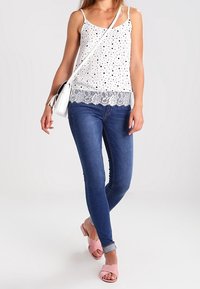Top de tirantes blanco con puntos negros y ribete de encaje, combinado con jeans skinny azules y sandalias de tacón rosas, con un pequeño bolso cruzado blanco.