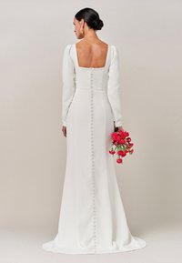 Abito bianco con maniche lunghe e schiena quadrata, caratterizzato da una fila di bottoni lungo la colonna vertebrale e una silhouette fluida e morbida. Bouquet di fiori rosa.