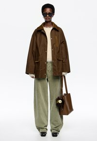 Hnedá nepremokavá bunda s čalúneným golierom, krémový sveter, olivovo zelené nohavice s širokými nohavicami a hnedá taška s dekoratívnym príveskom. Čierne slnečné okuliare.