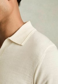 Cămașă polo de culoare crem, confecționată dintr-un material texturat, având un guler clasic și mâneci scurte, evidențiind detalii fine de cusătură.