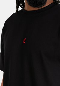 Zwarte katoenen t-shirt met een ronde hals en korte mouwen, voorzien van een klein rood geborduurd logo op het linkerbovenbeen.