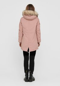 Parka rose avec une capuche doublée de fourrure, coupe droite et longueur mi-cuisse ; associée à un pantalon skinny noir et des bottes noires à lacets.