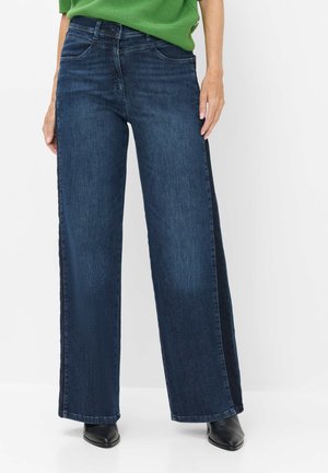 Person iført mørkeblå wide-leg jeans med en sort sidestribe, sorte spidse sko og en lysegrøn top mod en hvid baggrund.