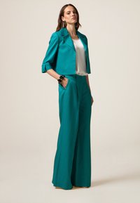 Blazer cropped color teal con maniche arrotolate abbinato a pantaloni palazzo a vita alta, dotati di tasche laterali e una texture liscia.