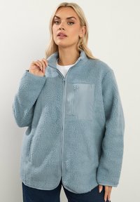 Giacca in pile azzurro chiaro con tasca sul petto con zip, superficie esterna testurizzata, colletto alto e vestibilità rilassata. Adatta per un abbigliamento casual.