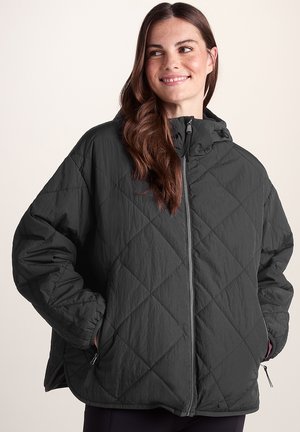 Smiling woman wearing a black quilted zip-up jacket with hood, hands in pockets, and long brown hair over one shoulder.