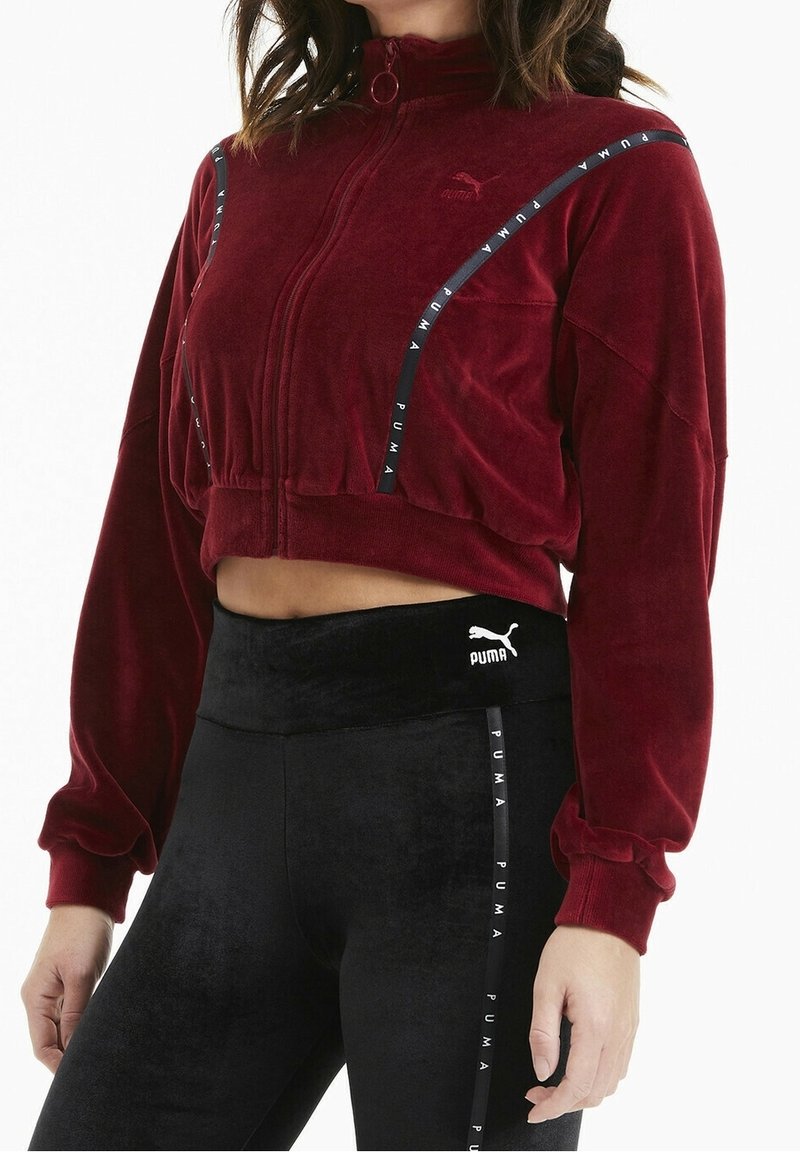 Femme portant une veste zippée courte Puma bordeaux et des leggings Puma noirs taille haute avec logo et bandes de marque.